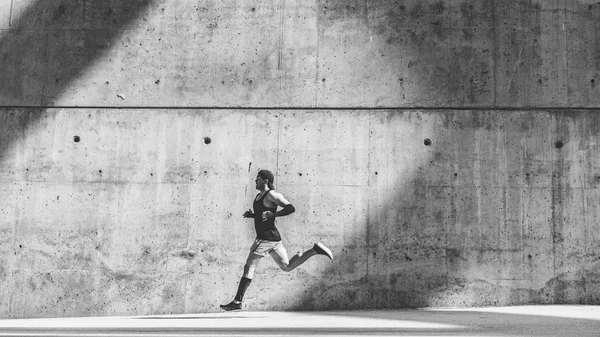 Hombre musculoso atleta velocista corriendo rápido, haciendo ejercicio al aire libre, trotando fuera contra fondo de hormigón gris con área de espacio de copia para el mensaje de texto o contenido del anuncio.Amplio, longitud completa.Blanco negro . —  Fotos de Stock