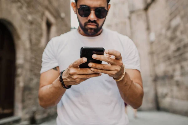 Young urban professional man using smart phone. Hipster coworker holding mobile smartphone using app texting sms message wearing sunglasses