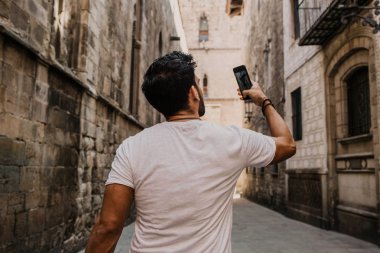 Beyaz tshirt kendini şehir seyahat tadını çıkarırken üstünde hareket eden telefon fotoğraf çekme içerik çekici İspanyol adam. Sakallı hippi erkek selfie smartphone üzerinden gotic sokaklarında yapma.