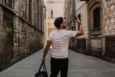 Beyaz tshirt kendini şehir seyahat tadını çıkarırken üstünde hareket eden telefon fotoğraf çekme içerik çekici İspanyol adam. Sakallı hippi erkek selfie smartphone üzerinden gotic sokaklarında yapma.