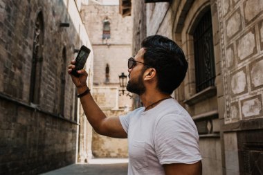 Beyaz tshirt kendini şehir seyahat tadını çıkarırken üstünde hareket eden telefon fotoğraf çekme içerik çekici İspanyol adam. Sakallı hippi erkek selfie smartphone üzerinden gotic sokaklarında yapma.