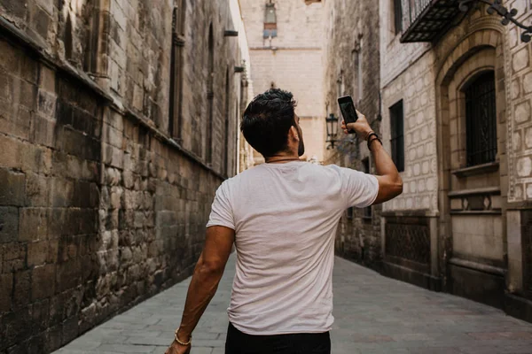 Contenido atractivo hombre hispano en camiseta blanca fotografiándose a sí mismo en el teléfono móvil mientras disfruta de los viajes de la ciudad.Varón hipster barbudo haciendo selfie a través de teléfono inteligente en las calles góticas . — Foto de Stock