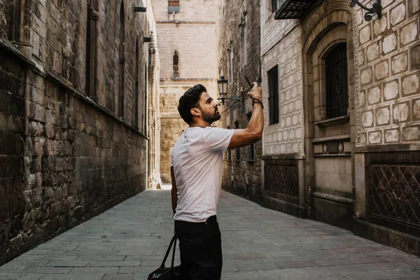 Contenido atractivo hombre hispano en camiseta blanca fotografiándose a sí mismo en el teléfono móvil mientras disfruta de los viajes de la ciudad.Varón hipster barbudo haciendo selfie a través de teléfono inteligente en las calles góticas . — Foto de Stock