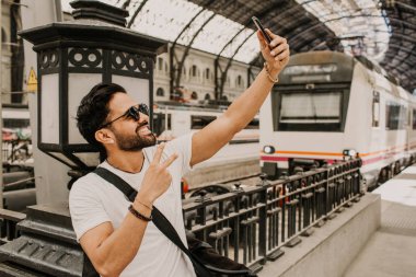 Beyaz tshirt kendini bekleyen tren Demiryolu istasyonu ise cep telefonuna fotoğraf çekme içerik çekici Hispanik erkek. Sakallı hippi erkek selfie smartphone üzerinden demiryolu platformu üzerinde yapma.