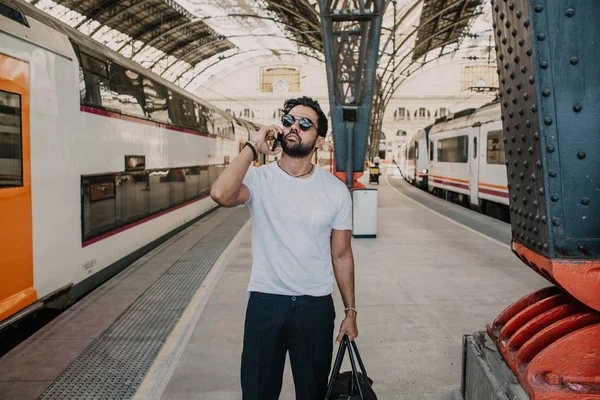Empresario barbudo positivo de confianza con gafas de sol de pie con bolsa de viaje en la sala de la estación de tren y el uso de teléfono móvil . — Foto de Stock