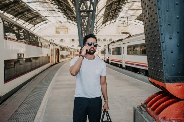 Empresario barbudo positivo de confianza con gafas de sol de pie con bolsa de viaje en la sala de la estación de tren y el uso de teléfono móvil . — Foto de Stock
