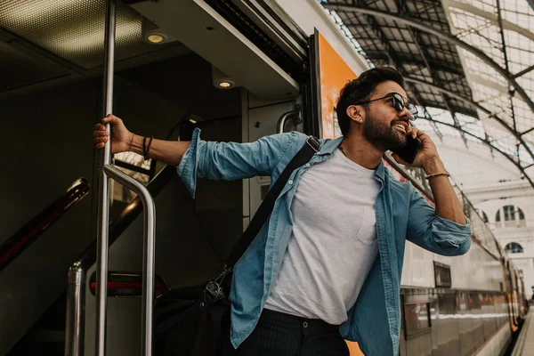 Confiado Atractivo joven hispano usando gafas de sol y camisa azul masculino sosteniendo la mano del teléfono inteligente y llamando a sus amigos mientras está parado en el vagón de tren en la plataforma — Foto de Stock