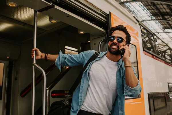 Atractivo joven hispano sonriente con gafas de sol y camisa azul hombre sosteniendo la mano del teléfono inteligente y llamando a sus amigos mientras está de pie en el vagón de tren en la plataforma — Foto de Stock