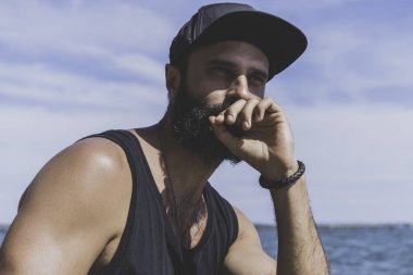 Sakallı adam modeli siyah kap ve tişörtü giyen, deniz sahne günbatımı görünüyor.