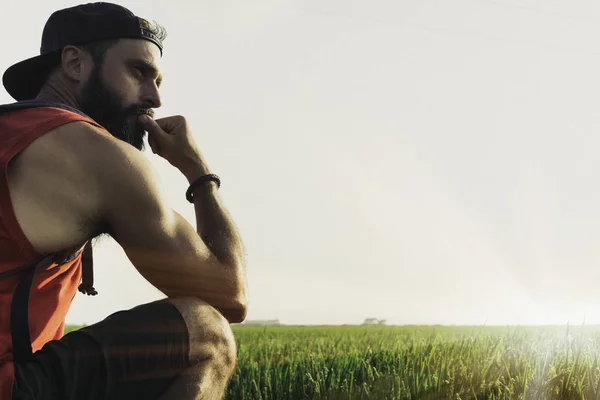 Modelo hombre barbudo con gorra negra y camiseta mira hacia otro lado, puesta de sol en el paisaje campos de arroz verde. Efectos visuales . —  Fotos de Stock