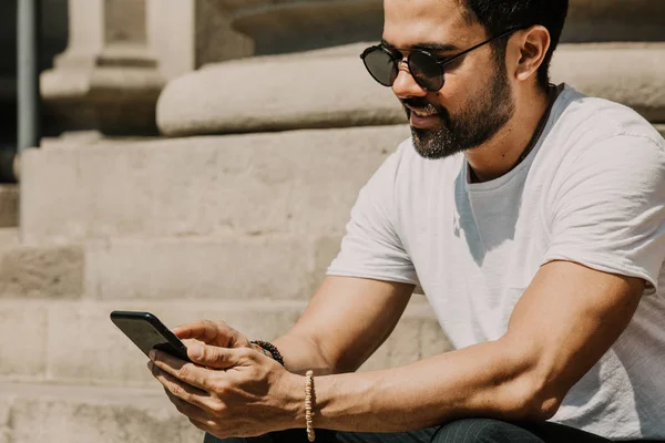 Молодой городской мужчина с помощью смартфона. Хипстер держит мобильный смартфон, используя смс с приложением в солнечных очках — стоковое фото