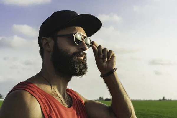 Bearded man model wearing black cap, sunglasses and red tshirt looks away, sunset in the green rice fields scenery