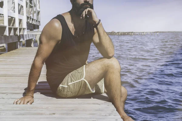 Pensativo hombre barbudo sentado en el muelle de madera en la orilla del mar y mirando a la distancia en el horizonte en el mar. concepto de emociones y sentimientos humanos. pensativo, anhelo y soledad —  Fotos de Stock