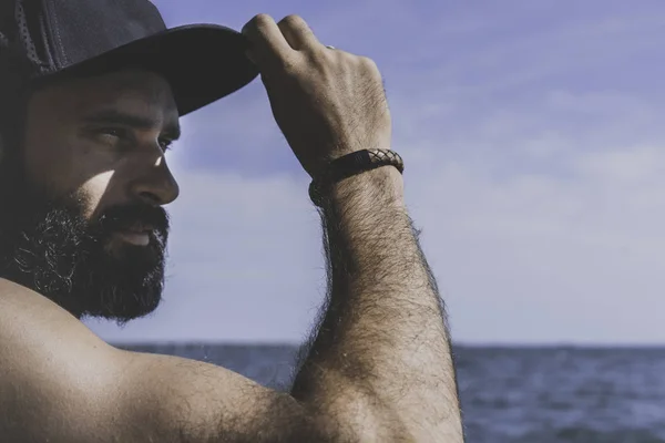 Retrato de cerca Modelo de hombre barbudo con gorra negra y camiseta mira hacia otro lado, puesta de sol en el paisaje del mar . —  Fotos de Stock