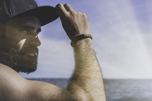 Modelo hombre barbudo con gorra negra y camiseta mira hacia otro lado, puesta de sol en el paisaje del mar. Efectos de luz solar . —  Fotos de Stock