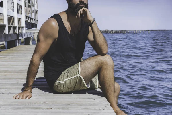 Pensativo hombre barbudo sentado en el muelle de madera en la orilla del mar y mirando a la distancia en el horizonte en el mar. concepto de emociones y sentimientos humanos. pensativo, anhelo y soledad —  Fotos de Stock