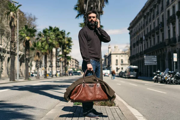 Atractivo hombre barbudo sosteniendo bolsa de viaje y el uso de teléfono móvil fuera. Fondo borroso . — Foto de Stock
