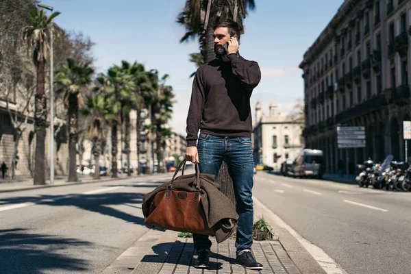 Atractivo hombre barbudo sosteniendo bolsa de viaje y el uso de teléfono móvil fuera. Fondo borroso . — Foto de Stock