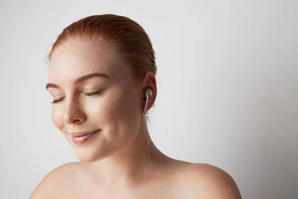 Retrato de encantadora pelirroja con sonrisa romántica y ojos cerrados escuchando música con auriculares inalámbricos en el oído sobre el fondo . — Foto de Stock