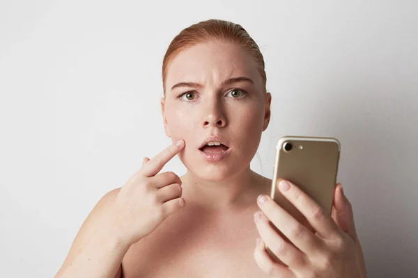 Kopf und Schultern einer rothaarigen Frau mit natürlichem Make-up. Enttäuschtes Mädchen blickt traurig auf Handy und zeigt mit dem Finger auf Pickel auf der Haut. — Stockfoto