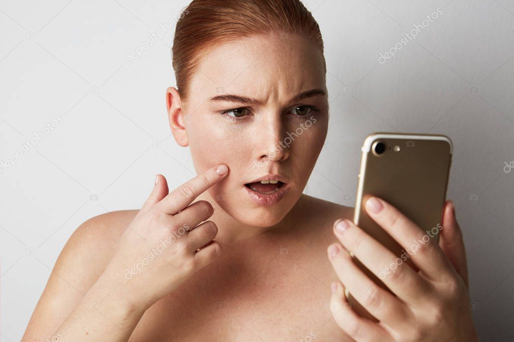 Head and shoulders of redhead woman with natural makeup. Disappointed girl looking at mobile phone sadly and pointing finger pimple on skin.