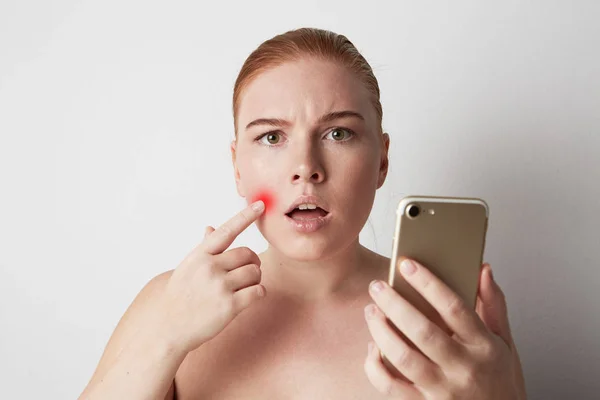 Kopf und Schultern einer rothaarigen Frau mit natürlichem Make-up. Enttäuschtes Mädchen blickt traurig auf Handy und zeigt mit dem Finger auf Pickel auf der Haut. — Stockfoto