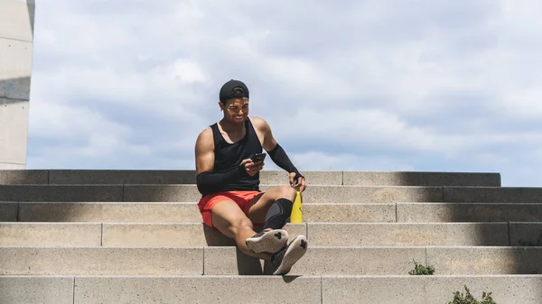 Sonriente deportista muscular ajuste hombre sprinter descansando después de su entrenamiento y el uso de teléfono móvil. Amplio —  Fotos de Stock