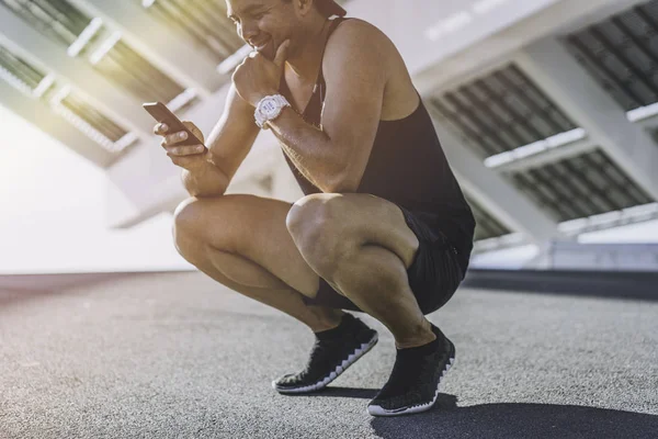 Sonriente ajustado muscular modelo deportivo velocista descansando después de su entrenamiento y el uso de teléfono móvil para comprobar sus resultados. Flarre. —  Fotos de Stock