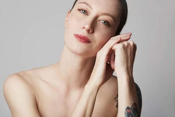 Mulher ruiva tatuada bonito com ombros nus mantém a mão perto do rosto sobre fundo cinza. Conceito de cosmetologia e cuidados com a pele . — Fotografia de Stock