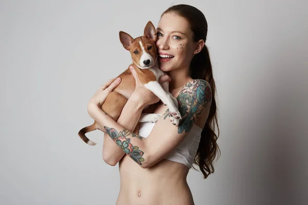 かわいいの肖像画は入れ墨ハグとキスを彼女の小さな子犬のバセニー犬の若い女性です。犬と所有者との間が大好きです。白い背景に分離. — ストック写真