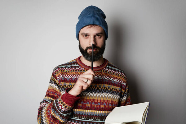 Handsome bearded man in blue beanie thinking with pen and notebook over background.