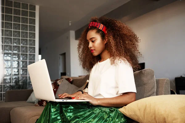 Jovem mulher africana americana digitando no laptop, enviando mensagens de texto para amigos através de redes sociais. Menina estudante navegando na Internet, usando wi-fi, sentado no sofá em casa . — Fotografia de Stock