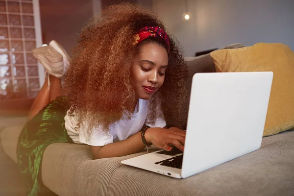 Mulher africana americana bonita digitando no laptop, mensagens de texto amigos através de redes sociais. Menina estudante navegando na Internet, usando wi-fi, sentado no sofá em casa . — Fotografia de Stock