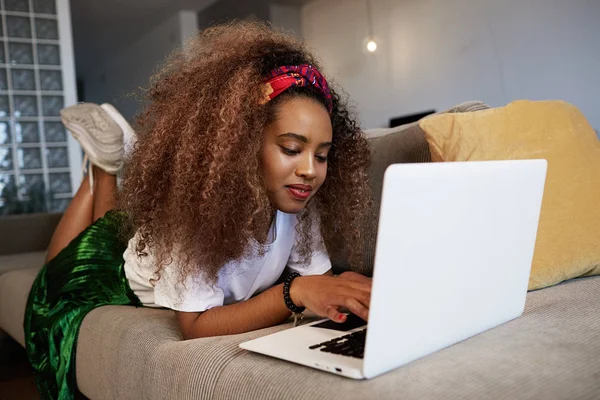 Cute american african woman typing on laptop, texting friends via social networks. Student girl browsing Internet, using wi-fi, sitting on sofa at home.