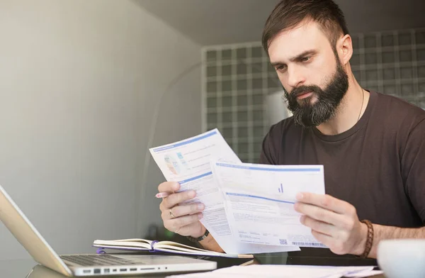 Bearded young man sitting at table looking at documents and thinking. Business man going through paperwork at home office