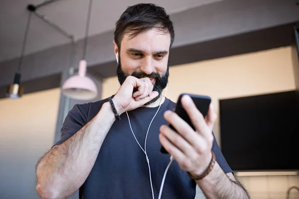 Красивый бородатый парень в обычной серой футболке, слушающий музыку в наушниках, проверяющий социальные сети на смартфоне, счастливый дома — стоковое фото