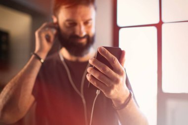 Akıllı telefonda sosyal ağlar kontrol, kulaklıkları rahat gri t-shirt dinleme müzik giyen emin sakallı adam evde mutlu