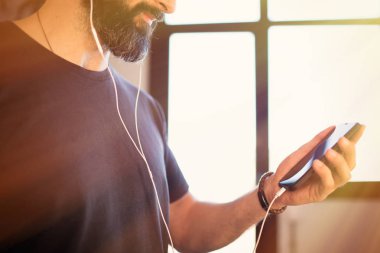 Gülümseyen sakallı adam kulaklıkla müzik dinleyen rahat gri t-shirt giyiyor, evde cep telefonu sosyal ağları kontrol
