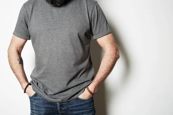 Atraente brutal tatuado barbudo cara posa em jeans azuis e branco cinza t-shirt premium verão algodão, no fundo branco mockup — Fotografia de Stock