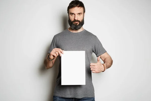 흰색 배경 모형에 빈 시트 종이와 빈 회색 티셔츠 프리미엄 여름 면화를 들고 긍정적 인 잔인 한 문신 수염 남자 — 스톡 사진