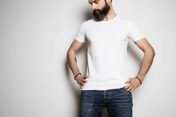 Handsome brutal tattooed bearded male model poses in blue jeans and blank white t-shirt premium summer cotton, on white background. — Stock Photo, Image