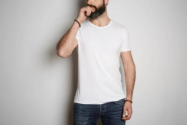 Jovem brutal tatuado barbudo cara posa em jeans azuis e branco branco t-shirt premium verão algodão, no fundo branco . — Fotografia de Stock