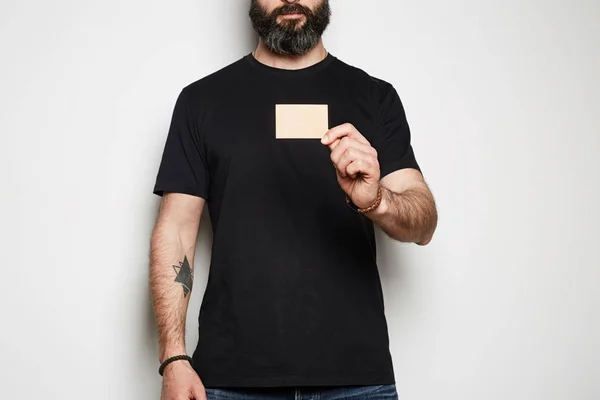 Güvenli sakallı hipster modeli rahat boş siyah t-shirt prim yaz pamuk pozlar ve beyaz arka planda el boş zanaat kartviziti tutarak. Mockup kopya yapıştırma alanı — Stok fotoğraf