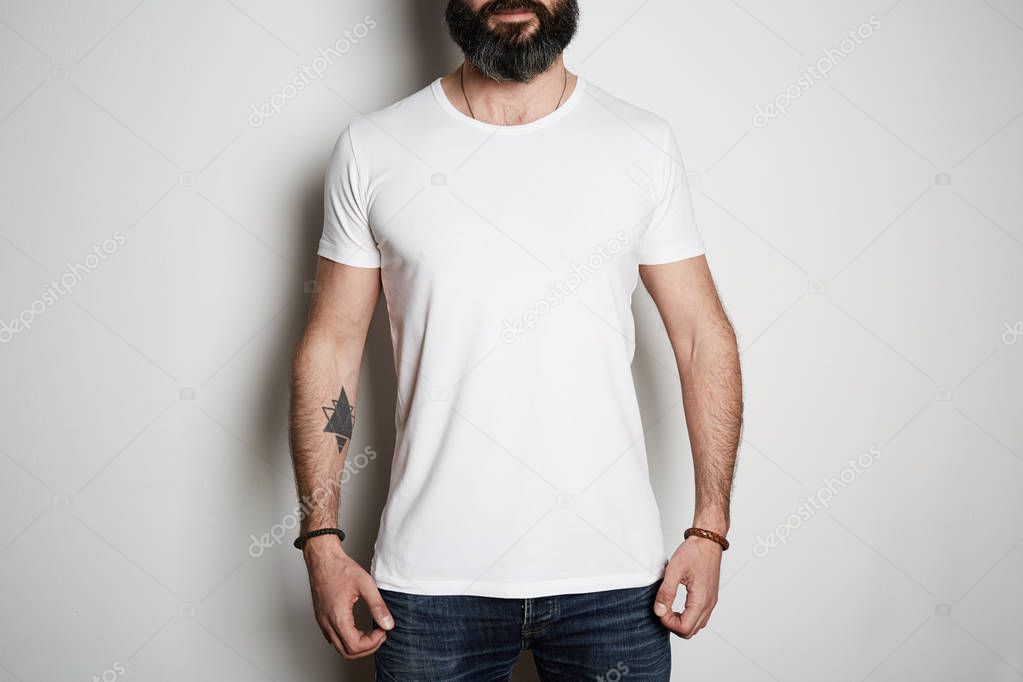 Attractive brutal tattooed bearded guy poses in blue jeans and blank gray t-shirt premium summer cotton, on white background mockup