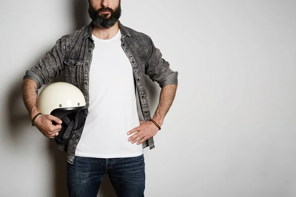 Attractive Brutal bearded male model poses in black jeans shirt and blank white t-shirt premium summer cotton with moto helmet in hands, on white background. Copy paste Advertisement.