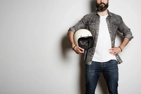 Trendy Brutal bearded male model poses in black jeans shirt and blank white t-shirt premium summer cotton with moto helmet in hands, on white background. Copy paste Advertisement. — Stock Photo, Image