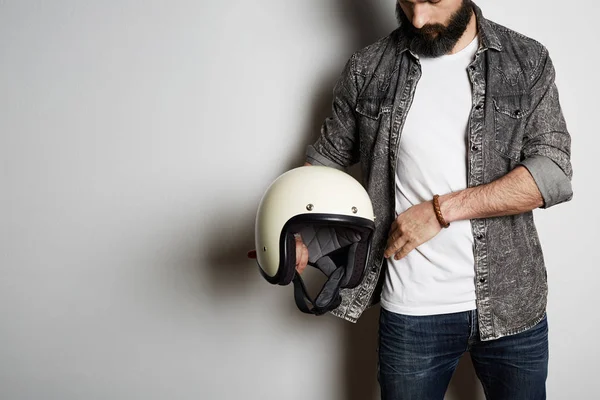 Stylish Brutal bearded male model poses in black jeans shirt and blank white t-shirt premium summer cotton with moto helmet in hands, on white background. Copy paste Advertisement. — Stock Photo, Image
