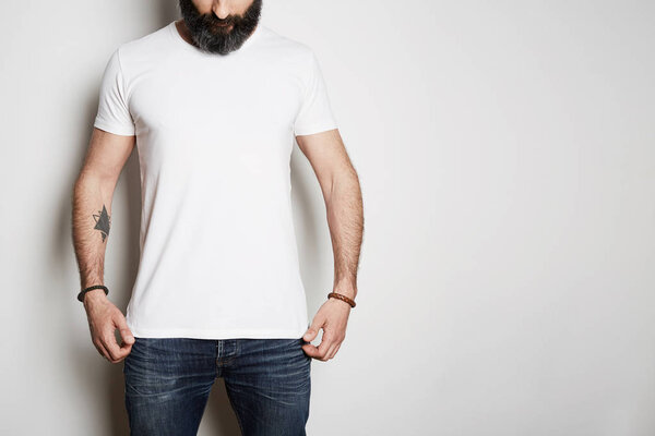 Handsome brutal tattooed bearded guy poses in blue jeans and blank gray t-shirt premium summer cotton, on white background. Mockup Copy Paste Advertisement.