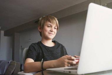 Evde oturma odasında masada otururken dizüstü bilgisayar kullanarak gülümseyen yakışıklı çocuk. Not defterine bakın.