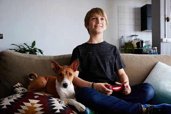 Happy cute boy playing video game console seated on a sofa with basenji dog puppy close in living room at home
